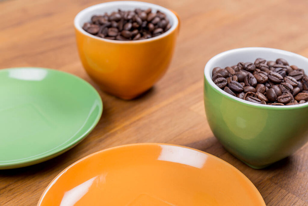 koffiesamenstelling, close-up van twee kopjes koffie een sinaasappel en een groene met hun borden vol koffiebonen op een houten ondergrond, horizontale foto - Foto, afbeelding
