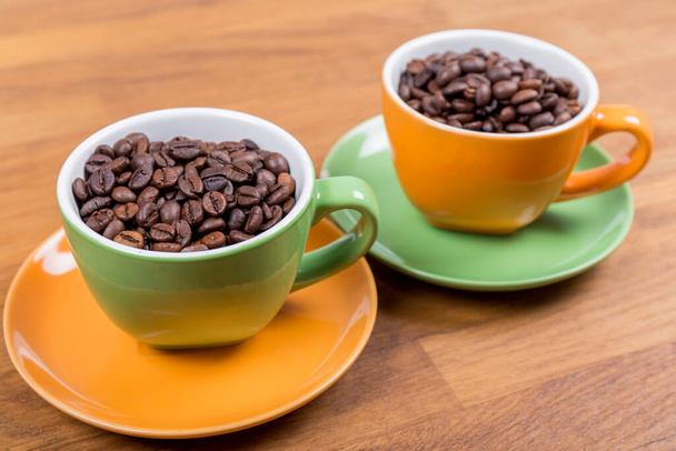 composition café, gros plan de deux tasses de café une orange et une verte avec leurs assiettes pleines de grains de café sur une surface en bois, photo horizontale - Photo, image