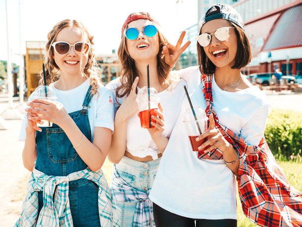Three young beautiful smiling hipster female in trendy summer clothes.Sexy carefree women posing outdoors.Positive models holding and drinking fresh cocktail smoothie drink in plastic cup with straw - Foto, Imagem