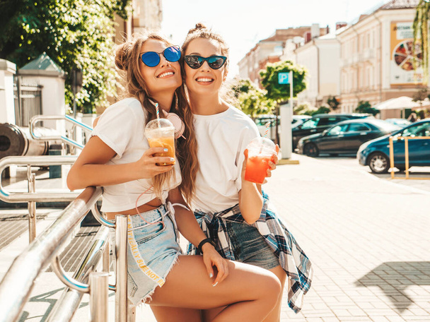 Two young beautiful smiling hipster female in trendy summer clothes. Carefree women posing outdoors.Positive models holding and drinking fresh cocktail smoothie drink in plastic cup with straw - 写真・画像