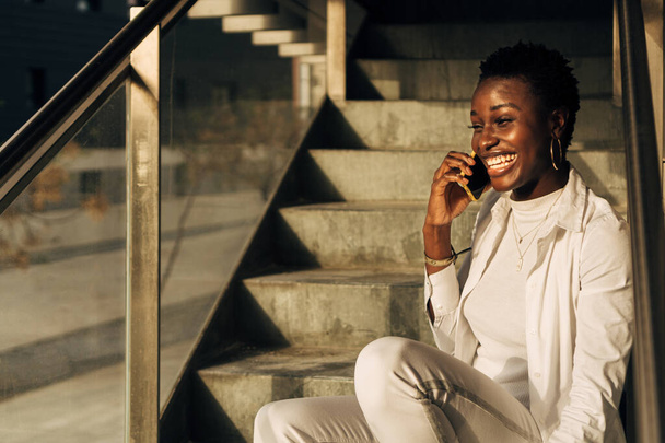 retrato de una chica africana de piel negra sentada en las escaleras y hablando por su teléfono celular - Foto, Imagen
