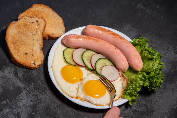 Deux œufs frits et des saucisses, laitue, concombre et tranches de radis sur une assiette blanche, pain grillé. Des œufs brouillés. Petit déjeuner. Une saine alimentation. Gros plan - Photo, image