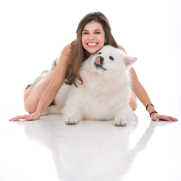 studiokuva nuoresta naisesta ja hänen valkoisesta koirastaan, istuen kädet ja polvet koiran päällä, katsoen eteenpäin, hymyillen ja koira katsoen sivulle, nuollen sen suuta. - Valokuva, kuva