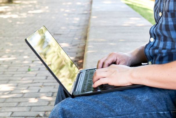 Nierozpoznawalny młodzieniec piszący na laptopie na zewnątrz. Koncepcja cyfrowego nomada, pracującego lub studiującego na świeżym powietrzu. - Zdjęcie, obraz