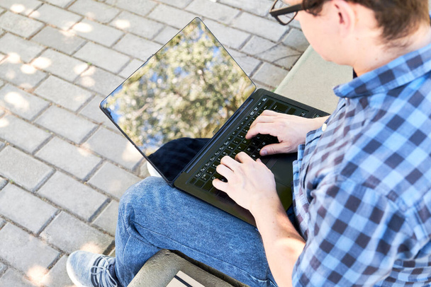 Młody człowiek pisze na laptopie na zewnątrz. Koncepcja cyfrowego nomada, pracującego lub studiującego na świeżym powietrzu. - Zdjęcie, obraz