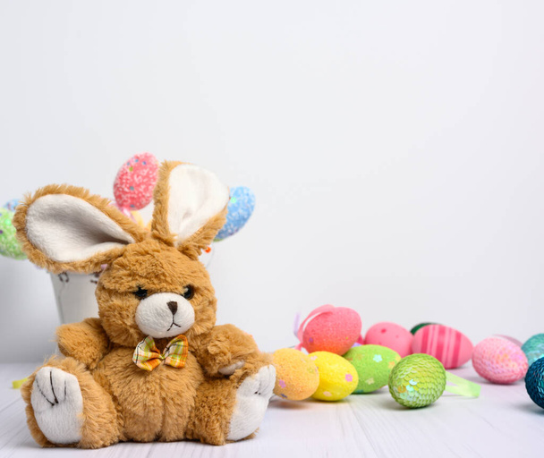 játék nyuszi ül egy fehér háttér és dekoratív színes húsvéti tojás, ünnepi háttér - Fotó, kép