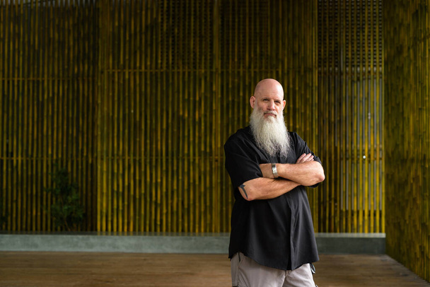 Portrait of confident man bald man with long gray beard outdoors against bamboo wall with arms crossed - 写真・画像