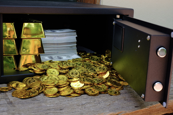 木製のテーブルの上にコインスタックと金の棒と紙幣100ドルの完全なスチールセーフボックス - 写真・画像