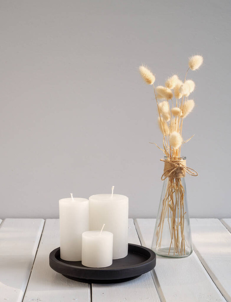 Білі свічки на чорному підносі і красиві лагурові сушені квіти в сучасній скляній вазі композиції над інтер'єром кімнати білого дерев'яного столу
 - Фото, зображення