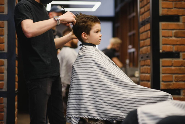 Side view of cute little boy getting haircut by hairdresser at the barbershop - Фото, изображение