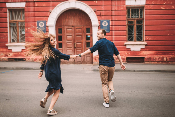A happy married couple in love runs down the street and rejoice. Beautiful young couple holding hands and smiling while walking along the city street - 写真・画像