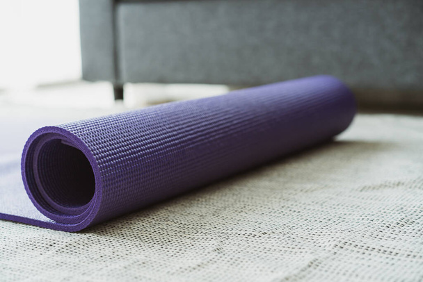 Rolled purple yoga exercise mat on the floor in living room close up with copyspace. Indoor workout activity concept. Rolling Purple Yoga carpet close up. - Φωτογραφία, εικόνα