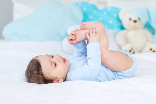 baby boy on the bed in a blue bodysuit playing with his feet, happy healthy little baby - Fotó, kép