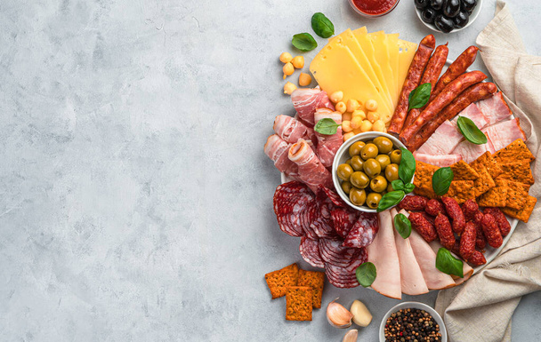 Meat and cheese snacks are sliced and laid out on a round board with olives and basil on a light gray background. Top view with copy space. - Fotó, kép