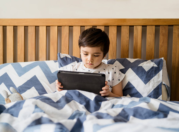 Kind liest vor dem Schlafengehen Bettgeschichten auf Tablet, Glücklicher Junge sitzt im Bett und spielt Spiele auf digitalem Pad, Kind entspannt am Wochenende zu Hause im Schlafzimmer. - Foto, Bild