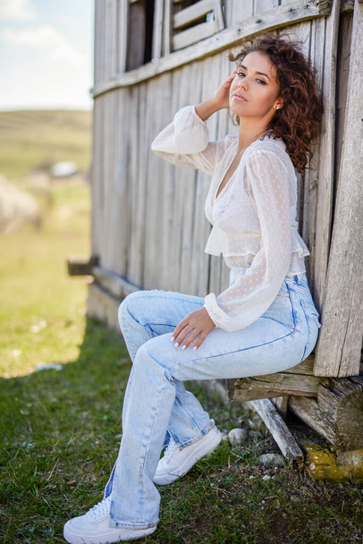 A beautiful woman with curly hair poses near an old wooden house. fashion style. - Foto, Bild