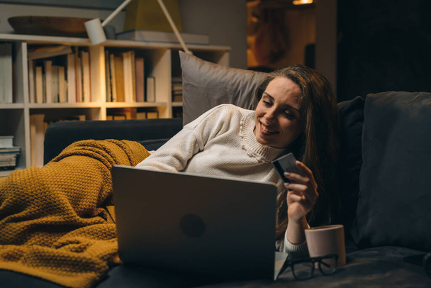 woman laying on sofa and shopping online using laptop computer at her home. evening atmosphere - Foto, Bild