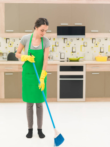 youn caucasian woman standing in kitchen, sweeping floor with broom - Photo, Image