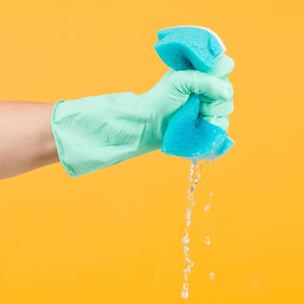 Hand in Schutzhandschuh quetscht Wasser aus Schwamm auf gelbem Hintergrund - Foto, Bild