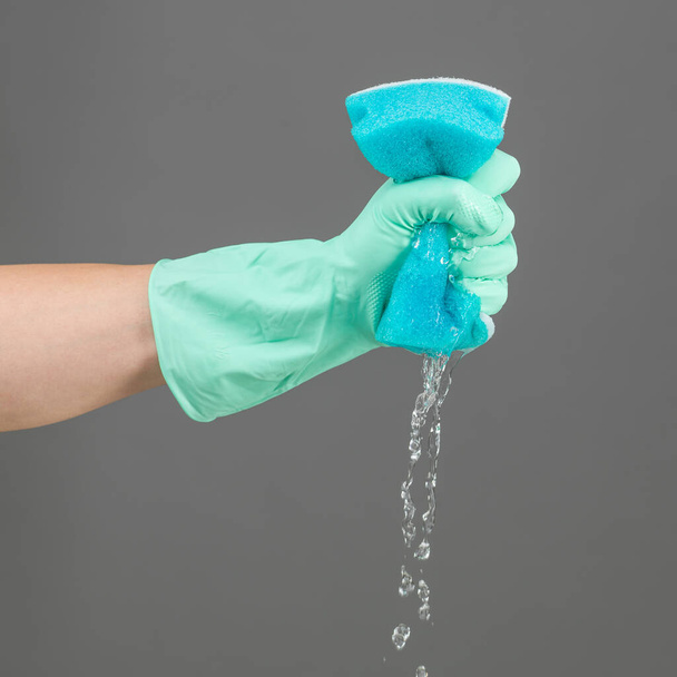 human hand with rubber glove squeezing water from sponge on grey background - Photo, Image