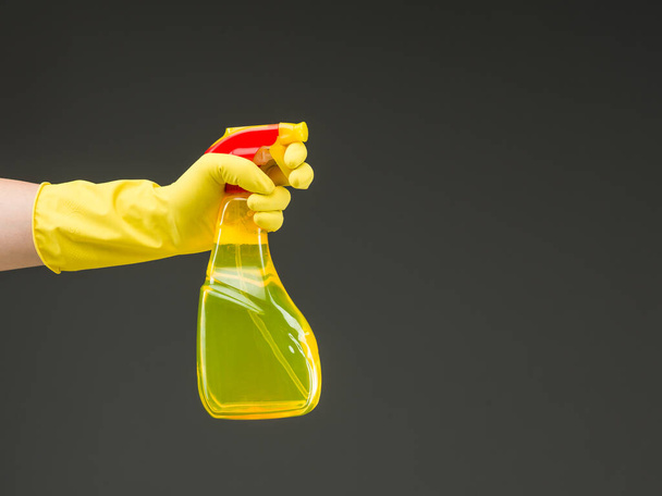mano con guante de goma que sostiene la botella del aerosol de la limpieza contra fondo gris - Foto, Imagen