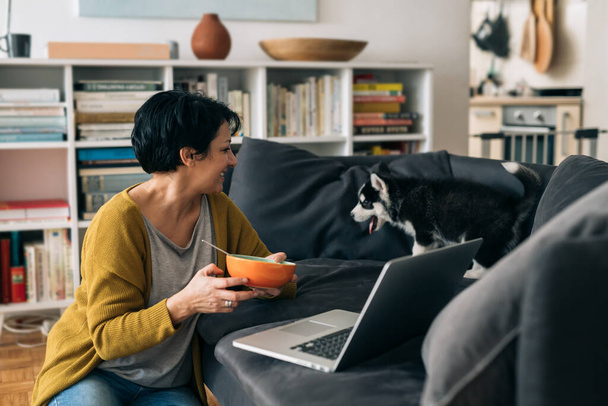 desayuno de la mujer en casa. está usando un ordenador portátil. su cachorrito está a su lado - Foto, Imagen