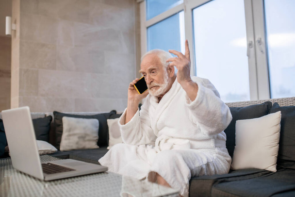 Gray-haired bearded man in white robe talking on the phone and looking excited - Fotoğraf, Görsel