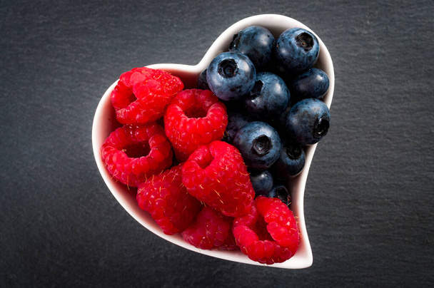 Heart healthy foods and good eating habits concept with a heart shaped white bowl of blueberries and raspberries spilt in the middle with a dark grey background - Photo, image