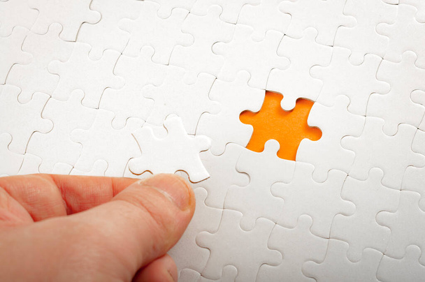 Completing final task, missing jigsaw puzzle pieces and business concept with hand holding the lacking white puzzle piece next to the incomplete or unfinished puzzle on orange background - Photo, Image