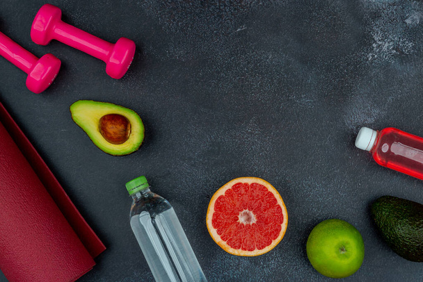Healthy lifestyle workout concept with training equipment. Pink dumbbells, yogamat, avocados, orange, grapefruit, and apple. Black cement background. Empty copy space. - Photo, Image