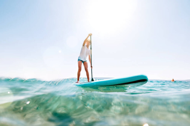 Under the water view angle to the smiling blonde teenager boy rowing stand up paddle board. Active family summer vacation time near the sea concept image. - Photo, image