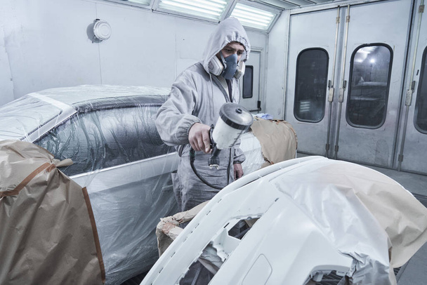 Car painting and automobile repair service. Auto mechanic in white overalls paints car with airbrush pulverizer in paint chambe - Фото, зображення