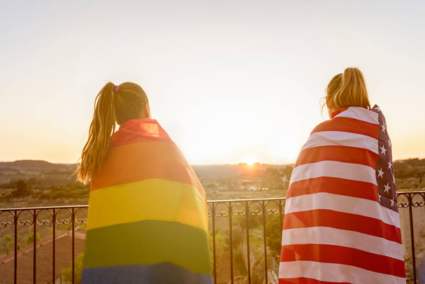 Dos mujeres jóvenes envueltas con la bandera del orgullo gay del arco iris y la bandera estadounidense al atardecer.. - Foto, imagen