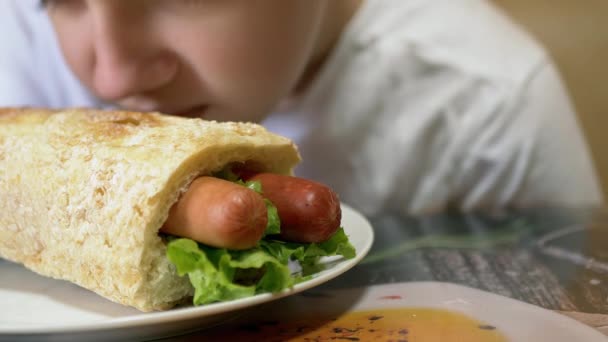 Hladové dítě čichá šťavnatý americký hot dog, olizuje rty s jazykem. 4K - Záběry, video