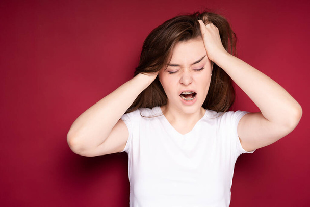 Pretty girl gets annoyed with headache after getting braces. Isolated on red background - Фото, изображение