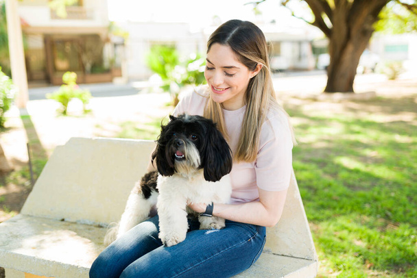 Mujer atractiva de unos 30 años sentada en un parque de bancos y sonriendo con un lindo perro en su regazo. Alegre amante del perro acariciando a un perro shih tzu  - Foto, Imagen