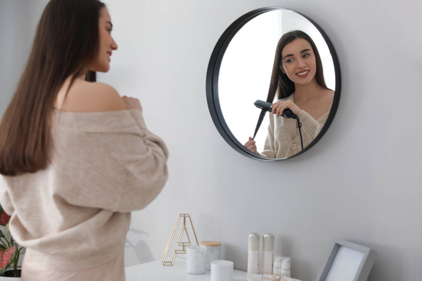 Young woman straightening hair near mirror at home. Morning routine - Foto, Imagem