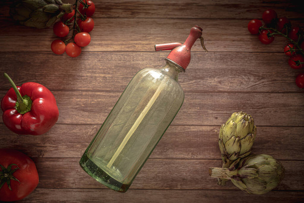 Flat lay retro food photography of an antique soda siphon, fresh cherry tomatoes, antique cutlery, artichokes and red pepper on a wood table. Still life image with a vintage look  - Фото, зображення