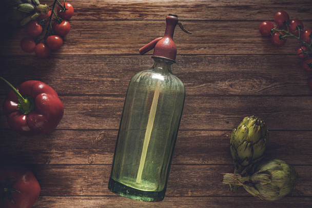 Flat lay retro food photography of an antique soda siphon, fresh cherry tomatoes, antique cutlery, artichokes and red pepper on a wood table. Still life image with a vintage look  - Foto, Bild