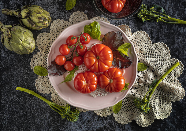 Moody dark food photography of fresh raf tomatoes, bimi and artichokes. Flat lay still life image with a vintage look and a black background. - Fotó, kép