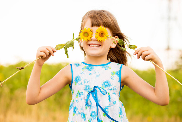 Roztomilé dítě dívka nosit šaty se slunečnicí v létě pole. Šťastná holčička skrývá oko se slunečnicí. Koncept letních prázdnin. - Fotografie, Obrázek