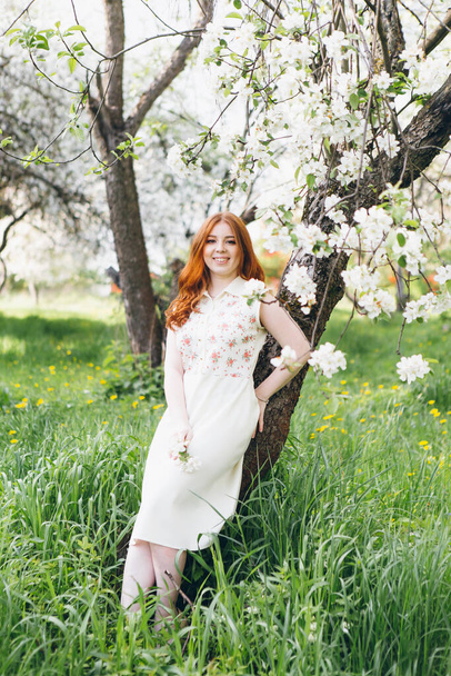 Young beautiful red-haired girl walks in a spring blooming apple orchard. Tinting. - Foto, Imagen