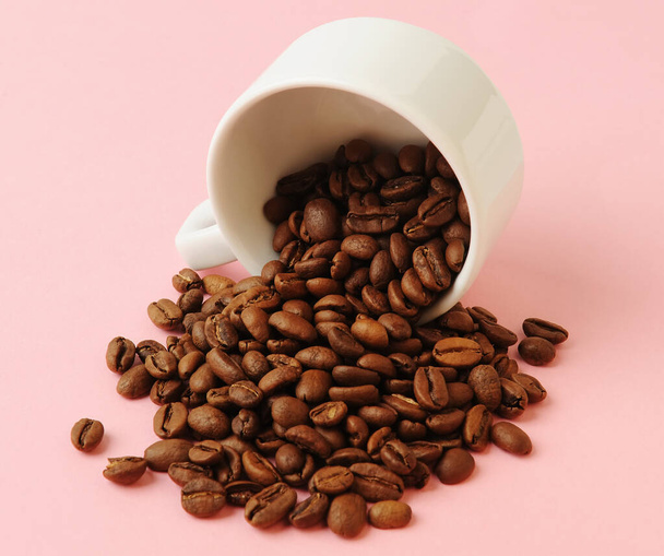 Omgekeerde witte beker met gegoten koffiebonen op zachte roze achtergrond close-up - Foto, afbeelding