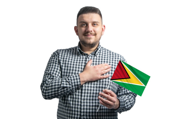 Hombre blanco sosteniendo una bandera de Guyana y sostiene su mano sobre su corazón aislado sobre un fondo blanco Con amor a Guyana. - Foto, imagen