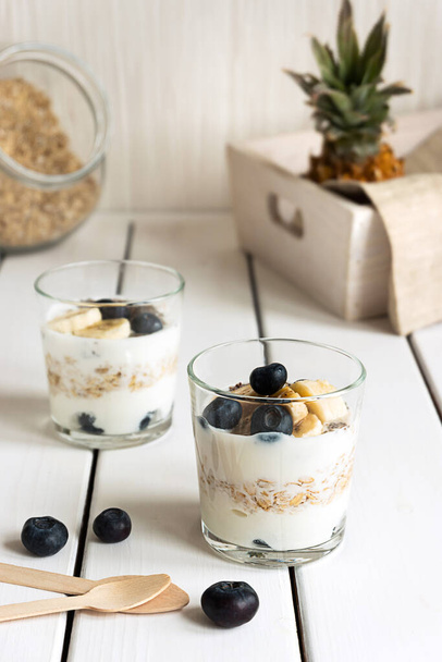 Dos vasos con yogur en capas, avena y frutas, sobre una mesa de madera blanca. Desayuno saludable, concepto de estilo de vida saludable - Foto, imagen