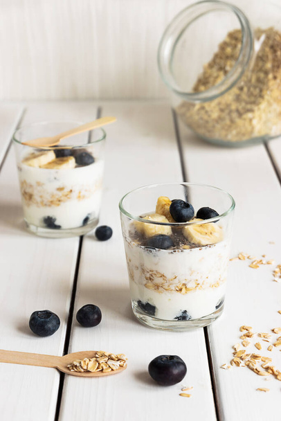 Dos vasos con yogur en capas, avena y frutas, sobre una mesa de madera blanca. Desayuno saludable, concepto de estilo de vida saludable - Foto, imagen