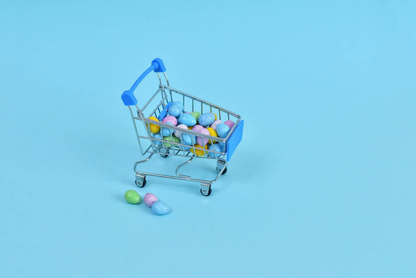 Carro de compras con huevos multicolores de Pascua sobre fondo azul. Venta de Pascua.Copiar espacio. Enfoque selectivo - Foto, Imagen