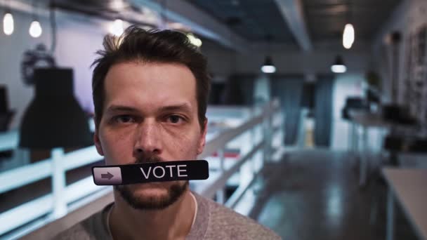 Мужчина смотрит в камеру и держит во рту табличку с надписью VOTE - Кадры, видео