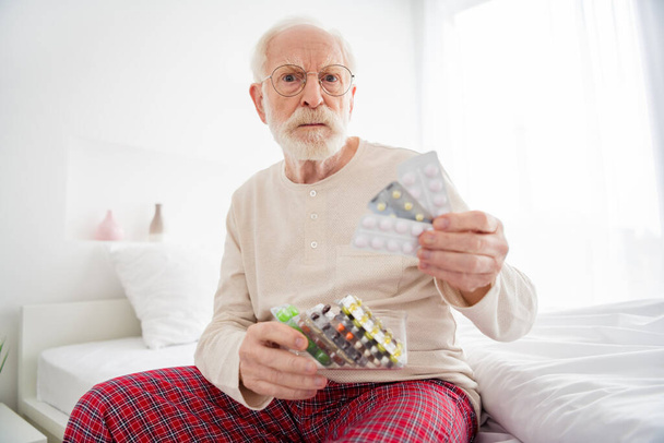 Photo of aged man pensioner unhappy sad unhealty unwell sit bed house look choose pills drugs painkiller - Φωτογραφία, εικόνα