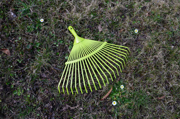 Un rastrello ventilatore giallo metallo si trova vicino a un palo di legno, e foglie autunnali sono sparsi su un prato con erba verde. rastrello ventilatore metallo sull'erba gialla in giardino. - Foto, immagini
